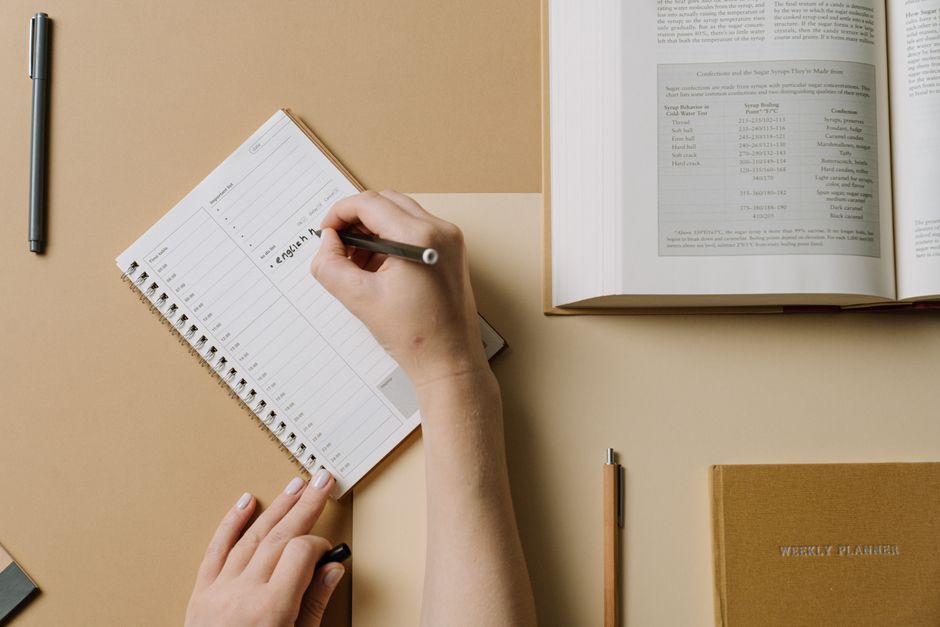 A person's hands writing in a planner at a desk with an open book nearby, conveying an organized workspace.